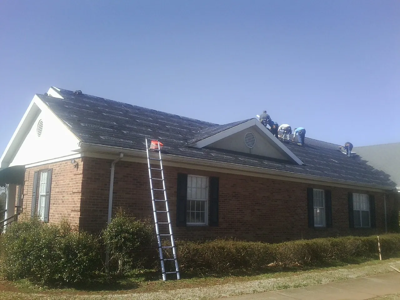 Crew tearing shingles off a brick ranch roof with a ladder set — Metal Roof Repair work by our crew in Lake Hill