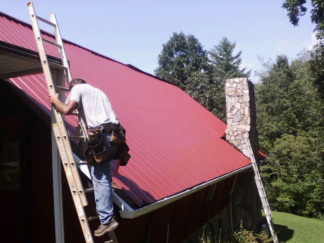 Roofer on a ladder beside a red metal roof and stone chimney — Soffit & Fascia Repair by Roofing Brothers in Lake Hill